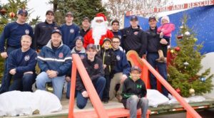 Caption: Lockwood Fire Chief John Staley posing with Santa along with his crew and volunteers at Canary and Bluebird in Lockwood during one of the visits. It takes a crew of about a dozen people to prep, help, take down and then move the Santa set from location to location. (Photo by Jonathan McNiven)
